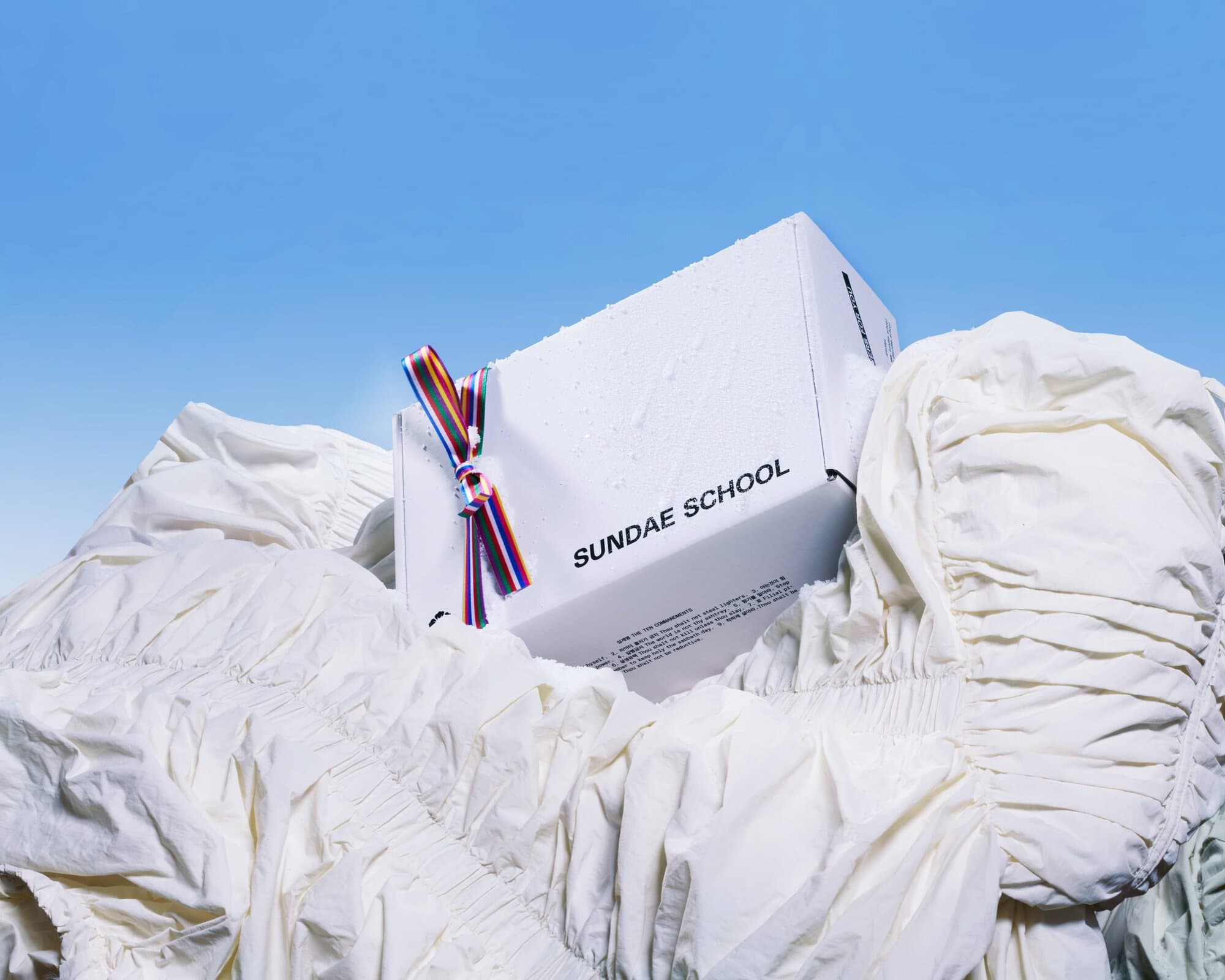 White Sundae School package tied with colorful striped ribbon resting among piled white fabric against a blue sky.