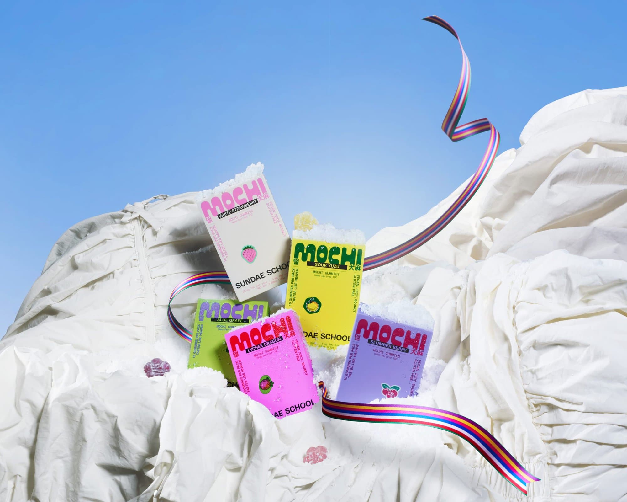 Colorful Mochi candy packaging arranged on draped white fabric under a bright blue sky.