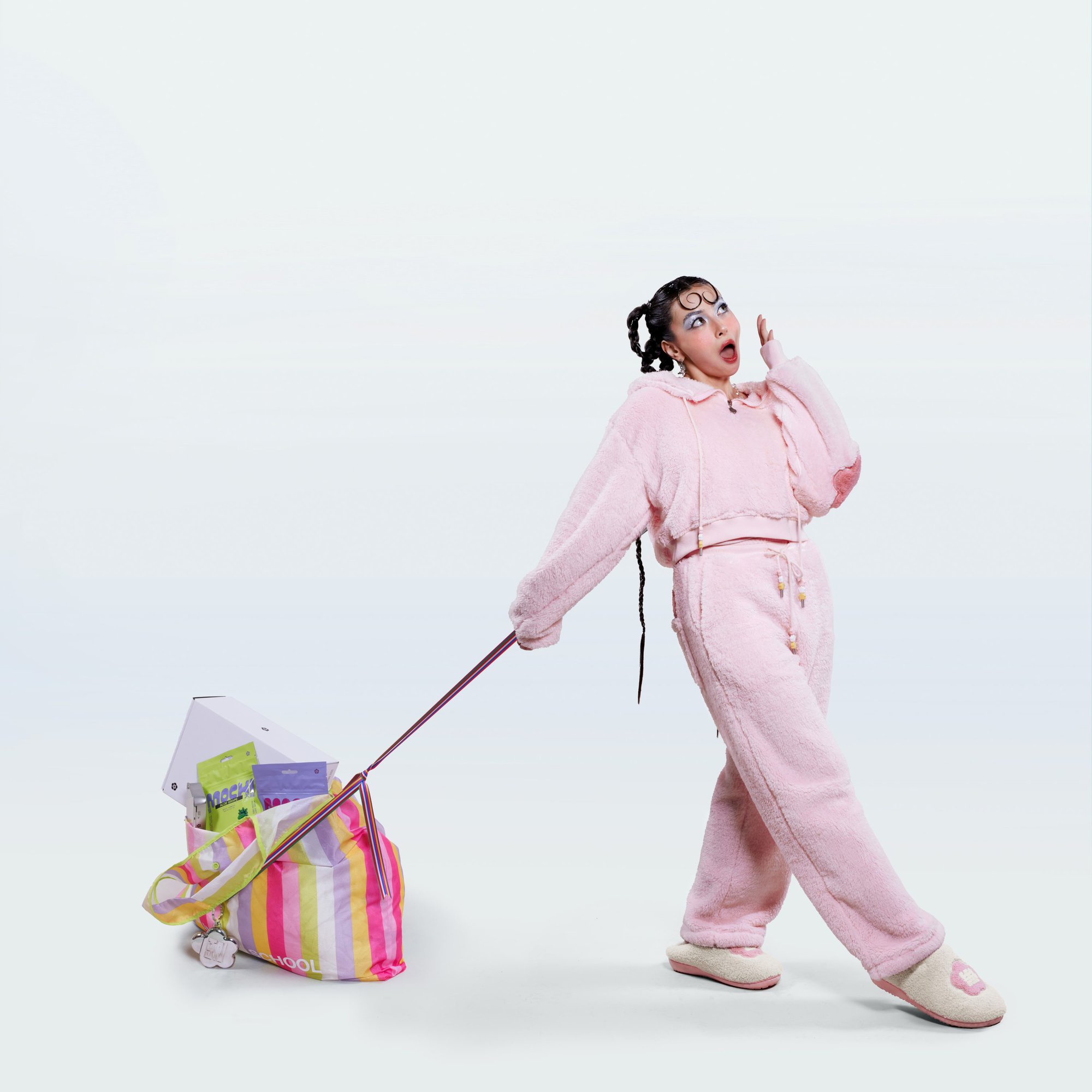 Model in pink fleece hoodie and matching pants pulling a striped bag on a white studio seamless background