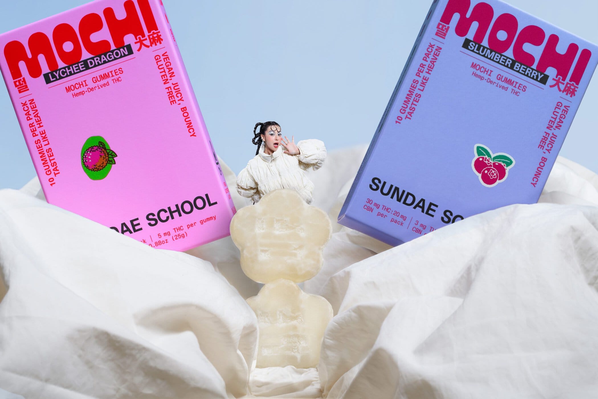 Model wearing a white puffer jacket standing between two large Mochi gummies product boxes by Sundae School against a pale background