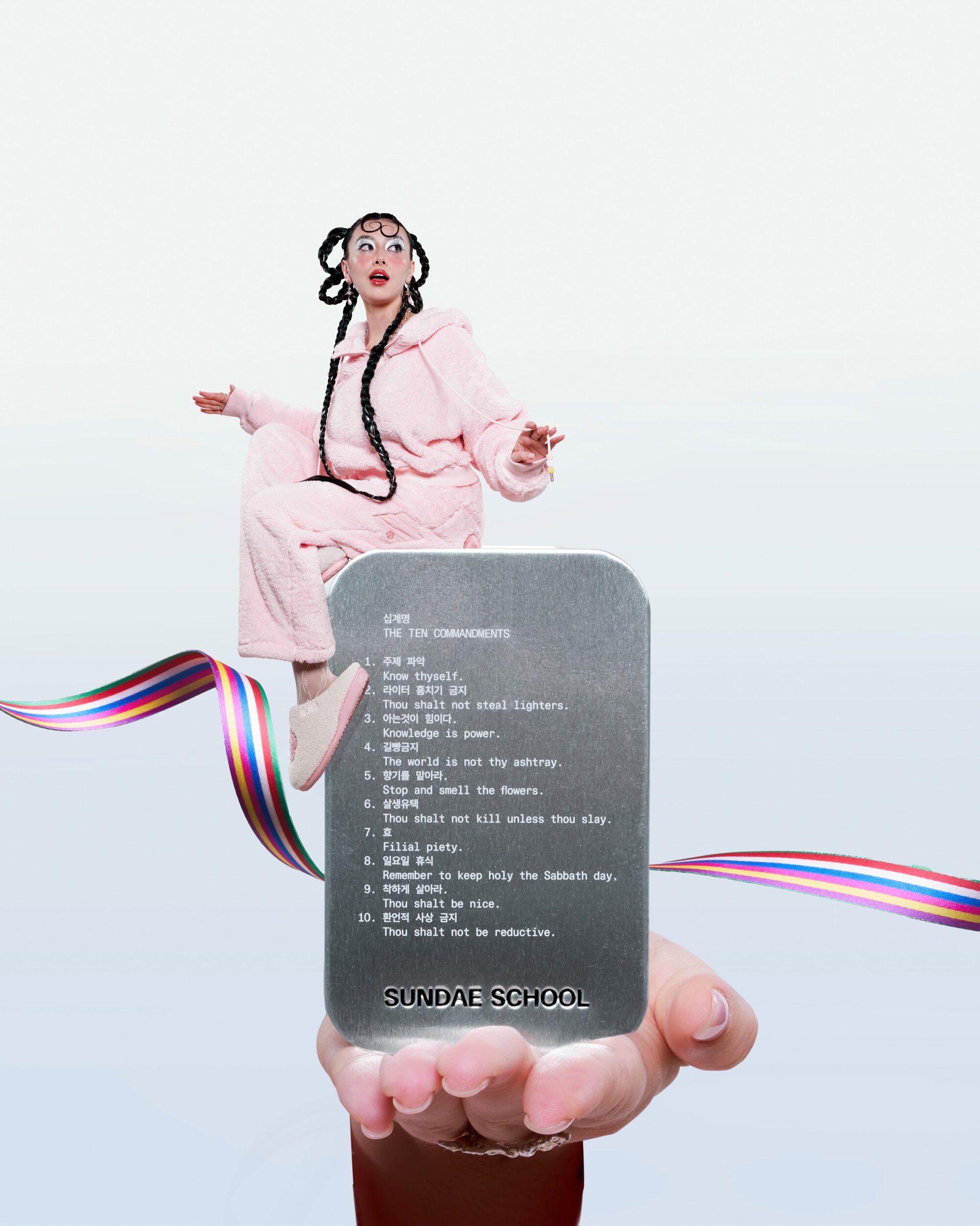 Model in pink hooded fleece sitting on a metallic tin held by a hand against a white backdrop with colorful ribbons.