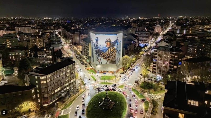 Aerial night view of a illuminated city traffic circle featuring a large Moncler Grenoble billboard ad on a building exterior.