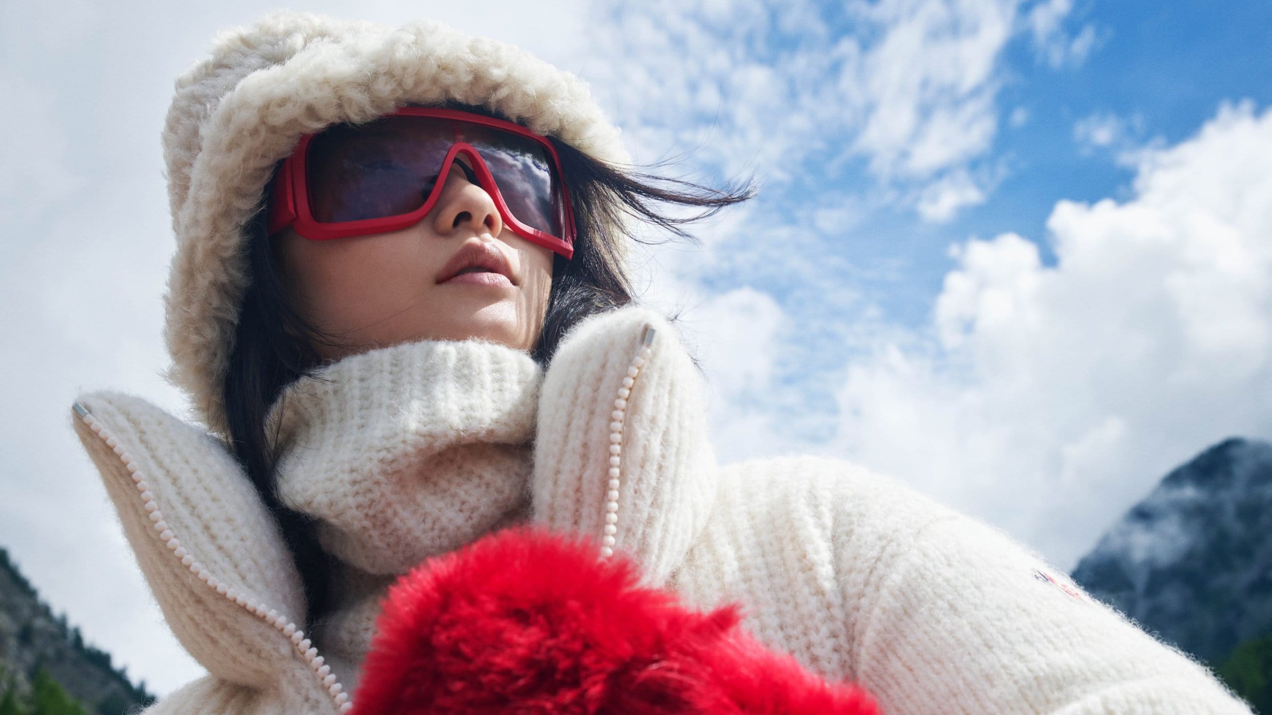 Model wearing white fuzzy hat, ski goggles, and white puffer jacket against a mountain sky background.