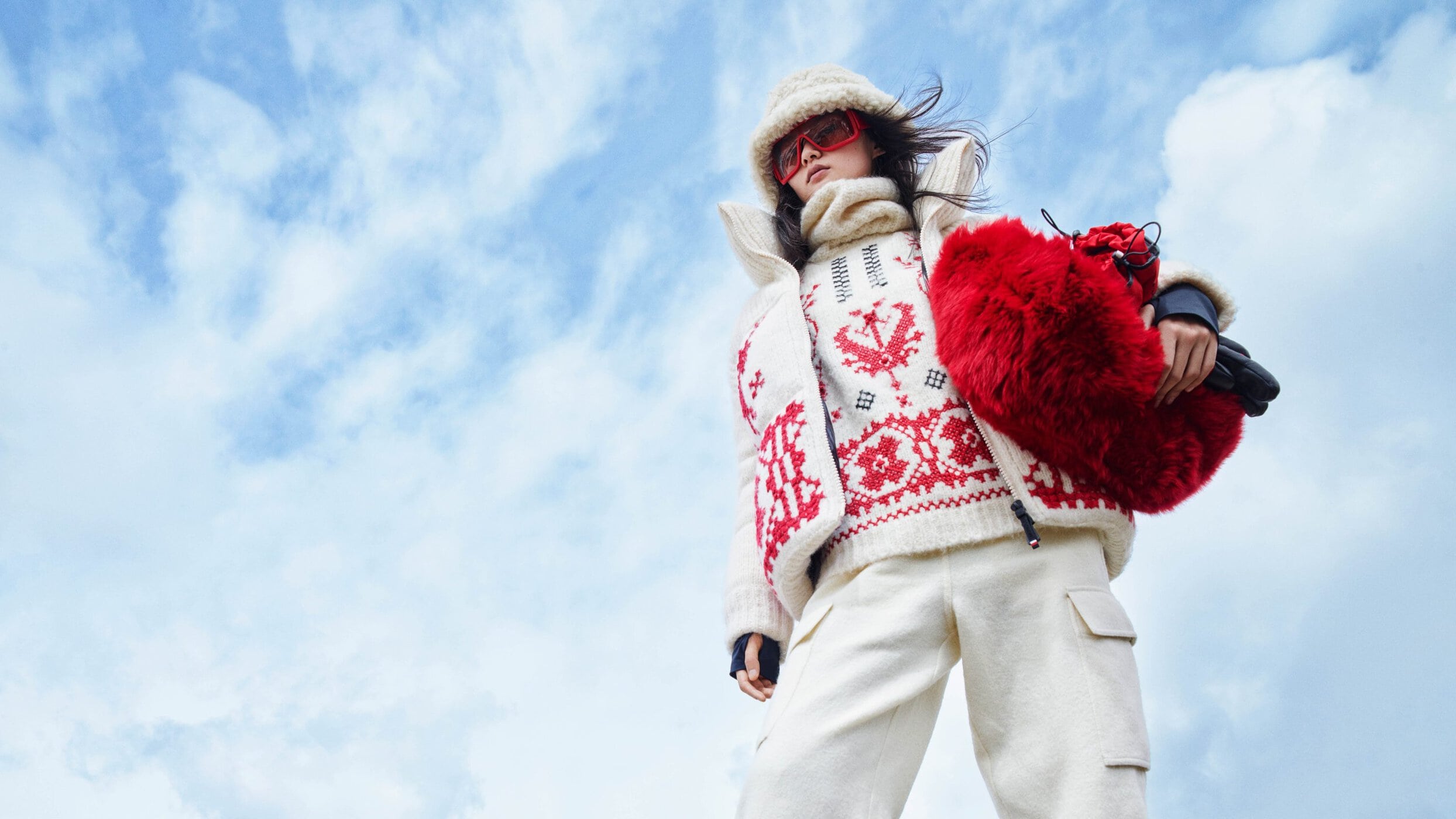 Model wearing a white patterned sweater, vest, and matching pants holding a bright red fuzzy bag against a blue sky background.