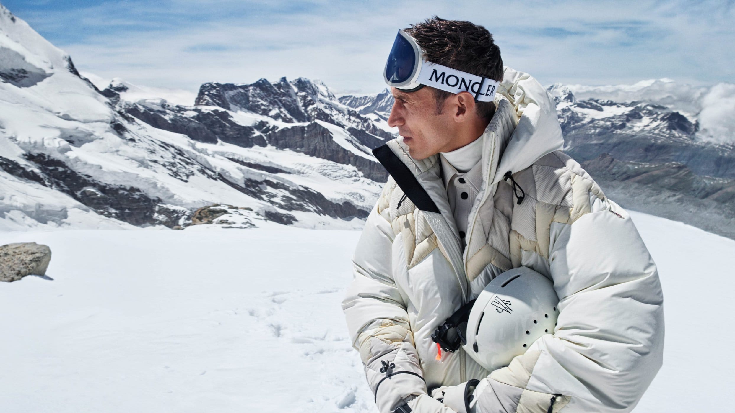 A man wearing white ski gear and goggles standing outdoors against a snowy mountain landscape