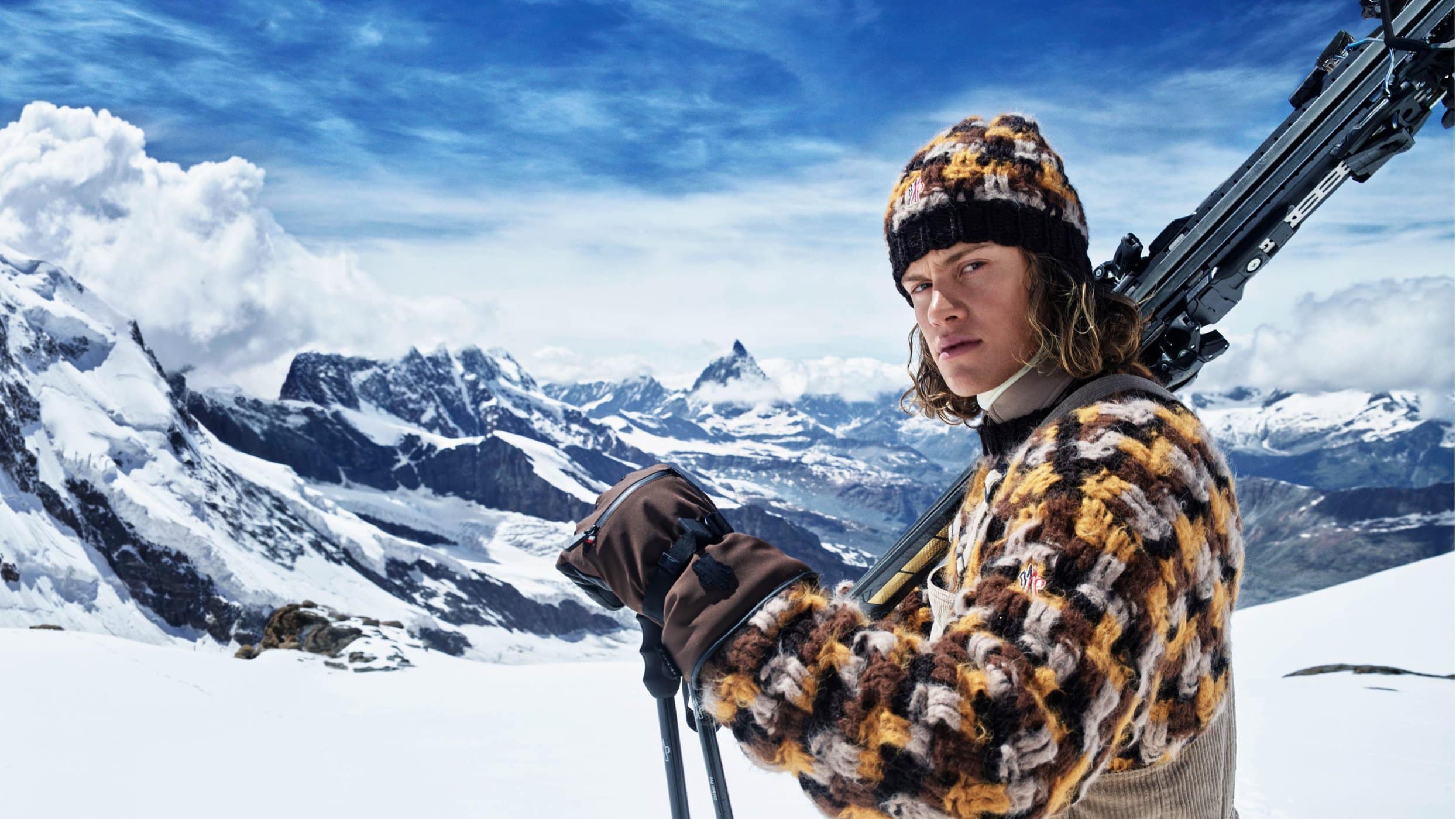 Man holding skis wearing a patterned knit hat and sweater against a snowy mountain range
