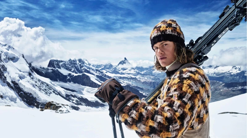 Man holding skis wearing a patterned knit hat and sweater against a snowy mountain range
