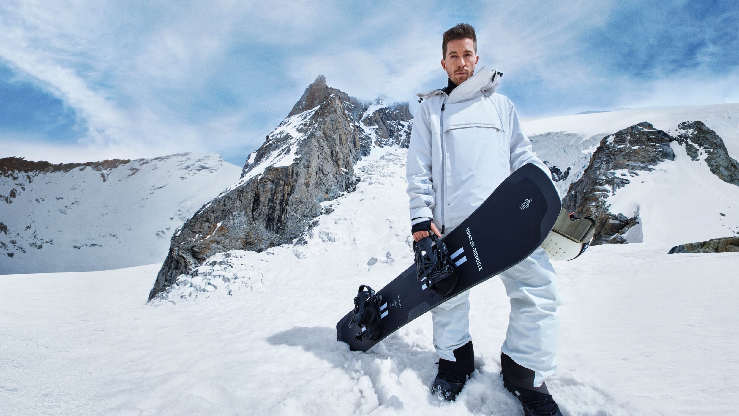 Shaun White holding a snowboard in a white ski suit against a snowy mountain landscape