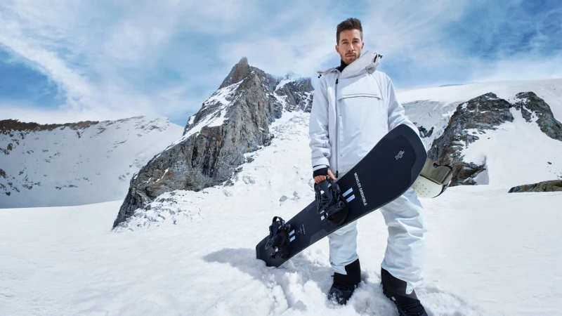 Shaun White holding a snowboard in a white ski suit against a snowy mountain landscape