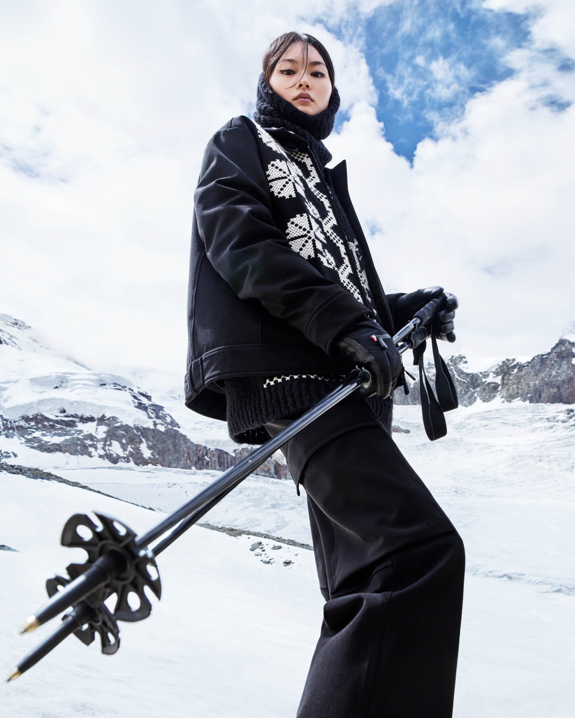 Model wearing black ski jacket and trousers holding ski poles against snowy mountain landscape under a partly cloudy sky.