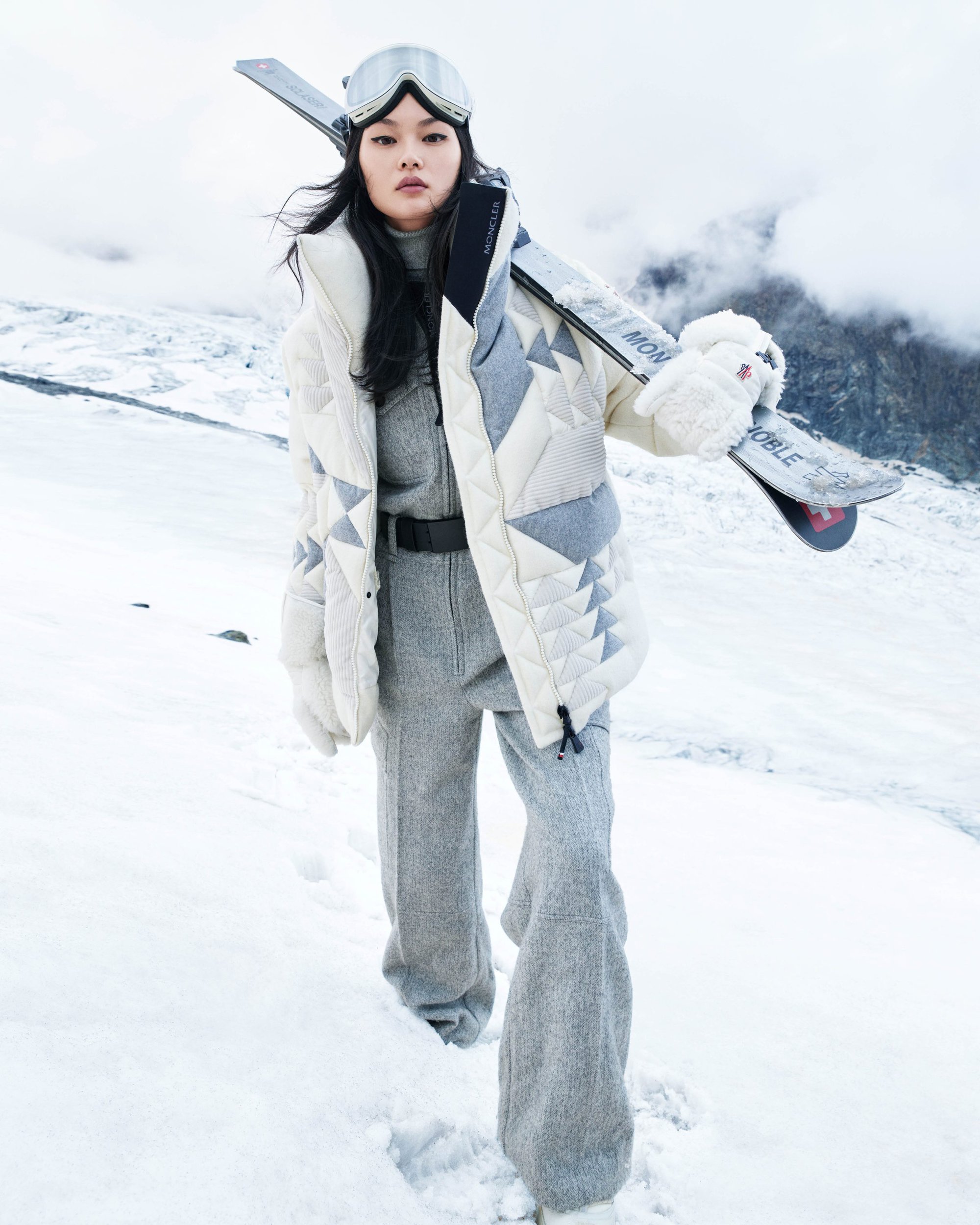 Model carrying skis wearing white winter jacket and grey trousers in a snowy mountain landscape