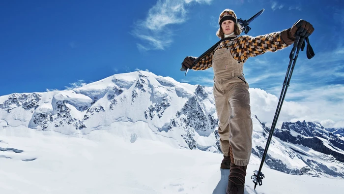 A skier in corduroy overalls and a patterned beanie stands on a snowy mountain holding ski poles, photographed by Mario Sorrenti.