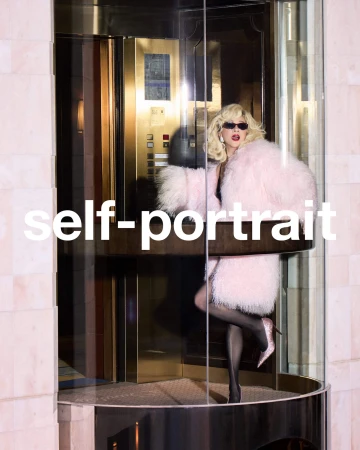 Blonde woman wearing a fluffy pink coat and sunglasses posing inside a glass elevator for Self-Portrait.