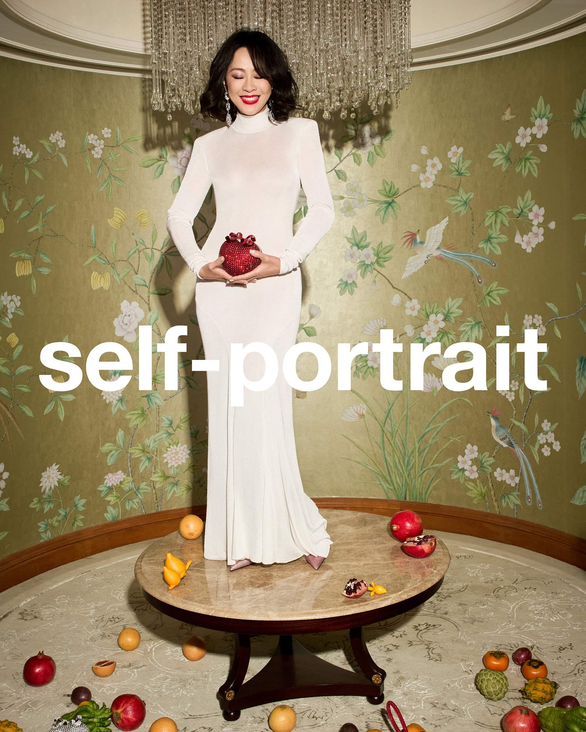Woman in a long white turtleneck dress holding a red sequined bag while standing on a round table decorated with fruit.