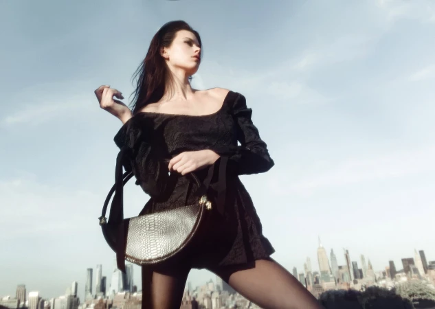 Model Georgia Fowler posing with a structured black handbag against an urban skyline under a pale sky.