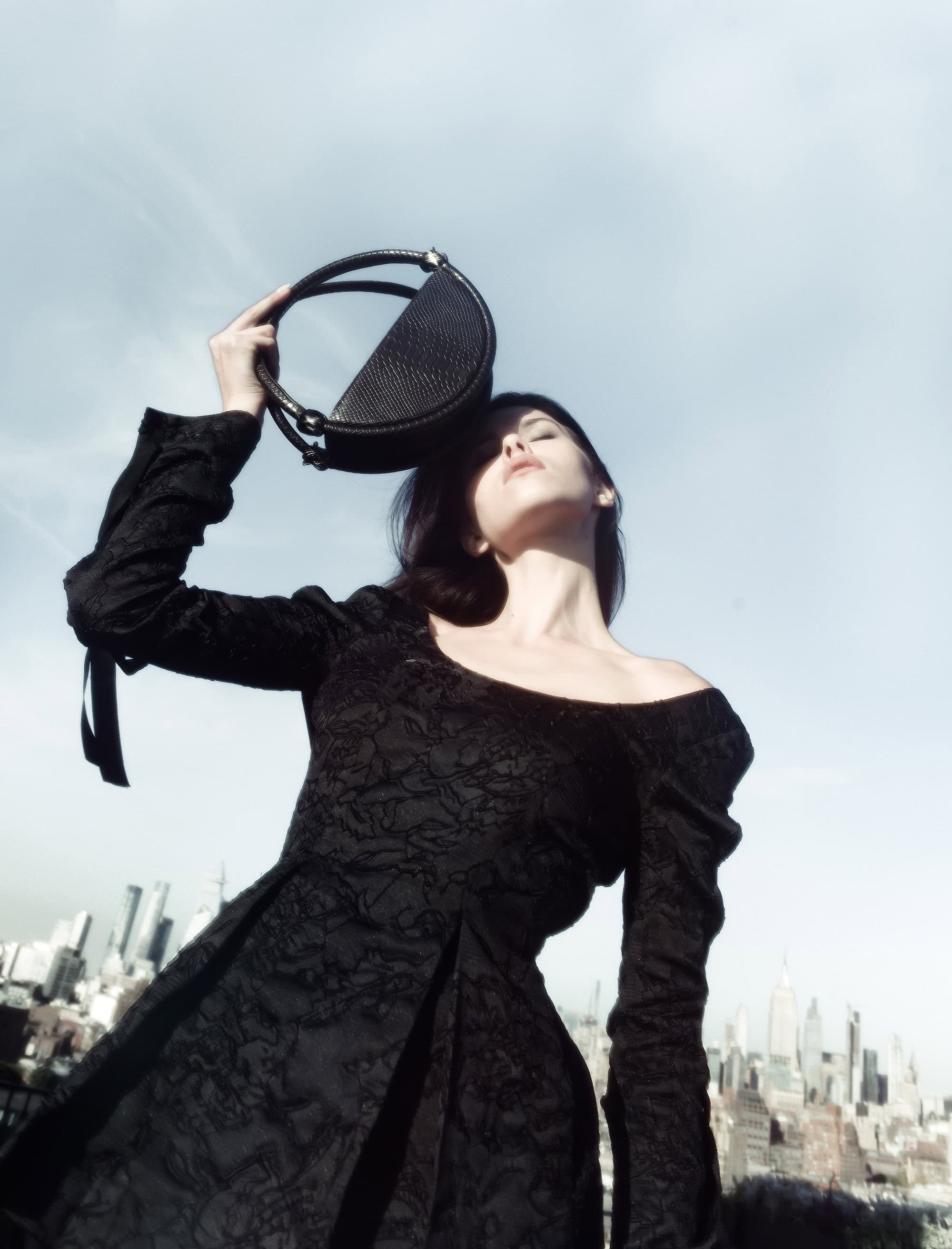 Model wearing black lace dress holding up a dark circular handbag against a city skyline under clear sky