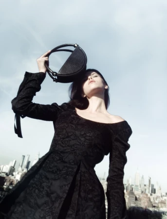 Model wearing black lace dress holding up a dark circular handbag against a city skyline under clear sky