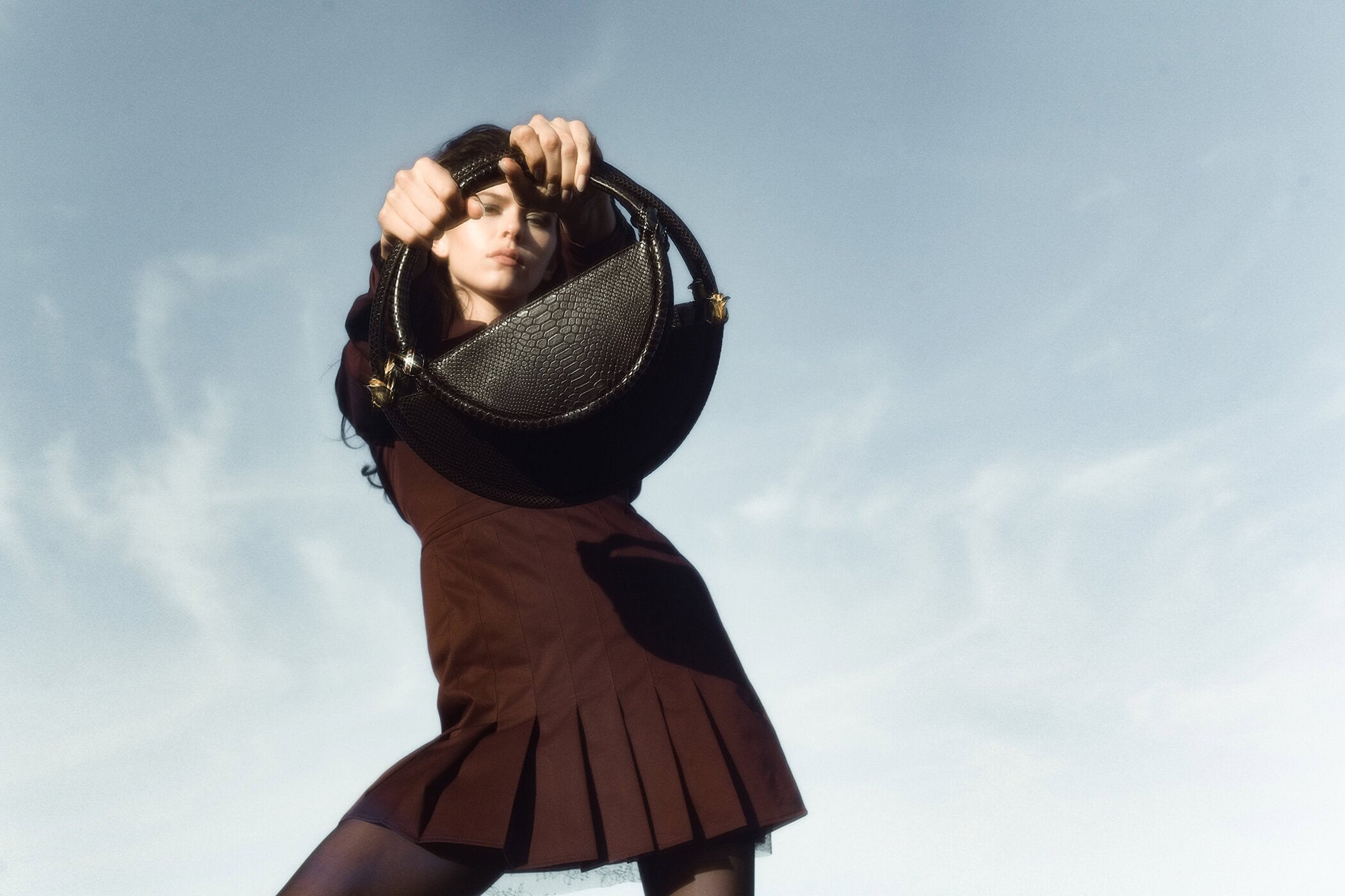 Model Georgia Fowler holding a black handbag in front of her face against a pale sky for Contessa Mills