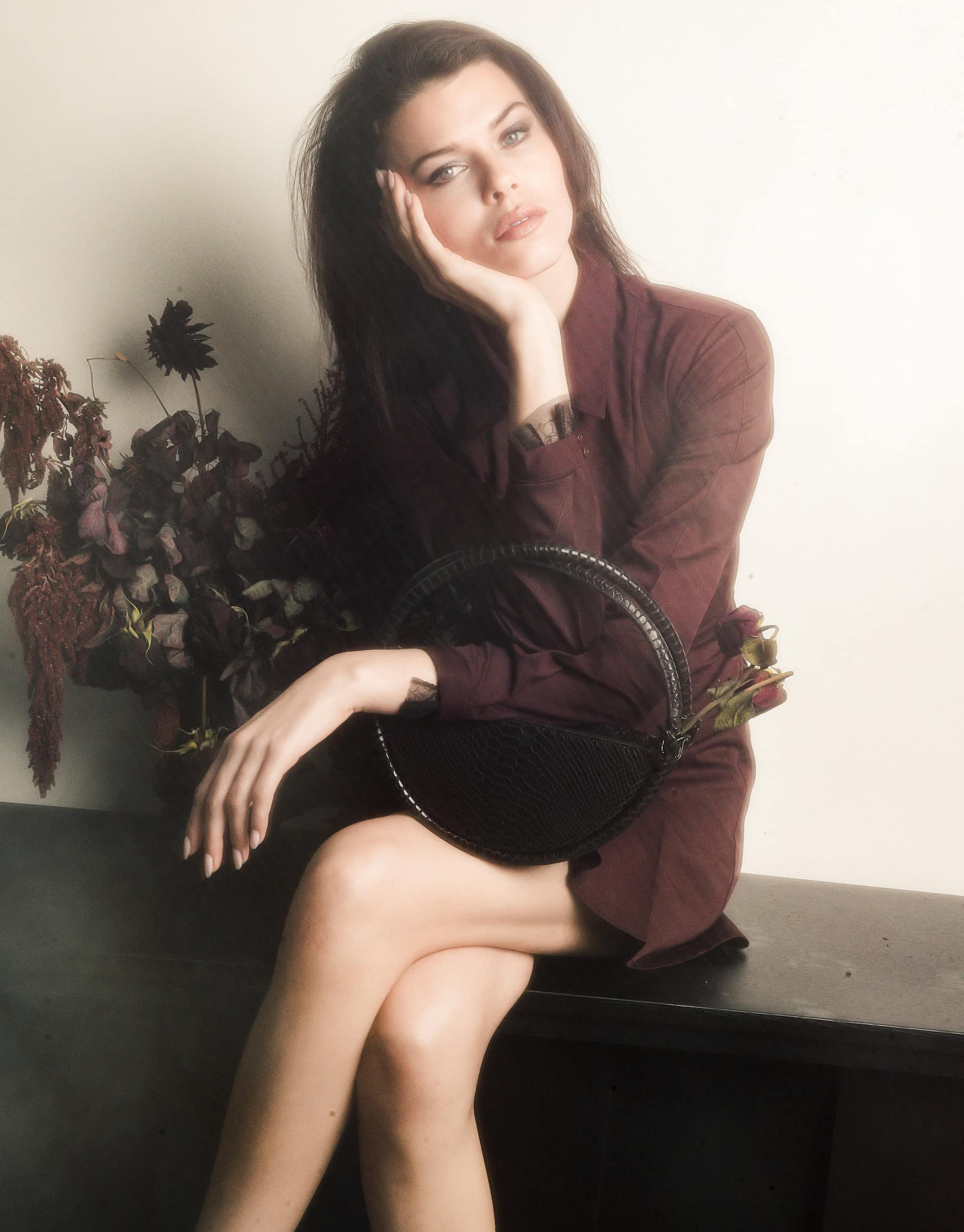 Model seated on a dark ledge holding a round black handbag next to dark floral arrangements against a plain wall.