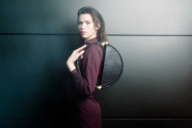Model wearing a dark purple dress holding a black circular handbag against a dark panelled wall