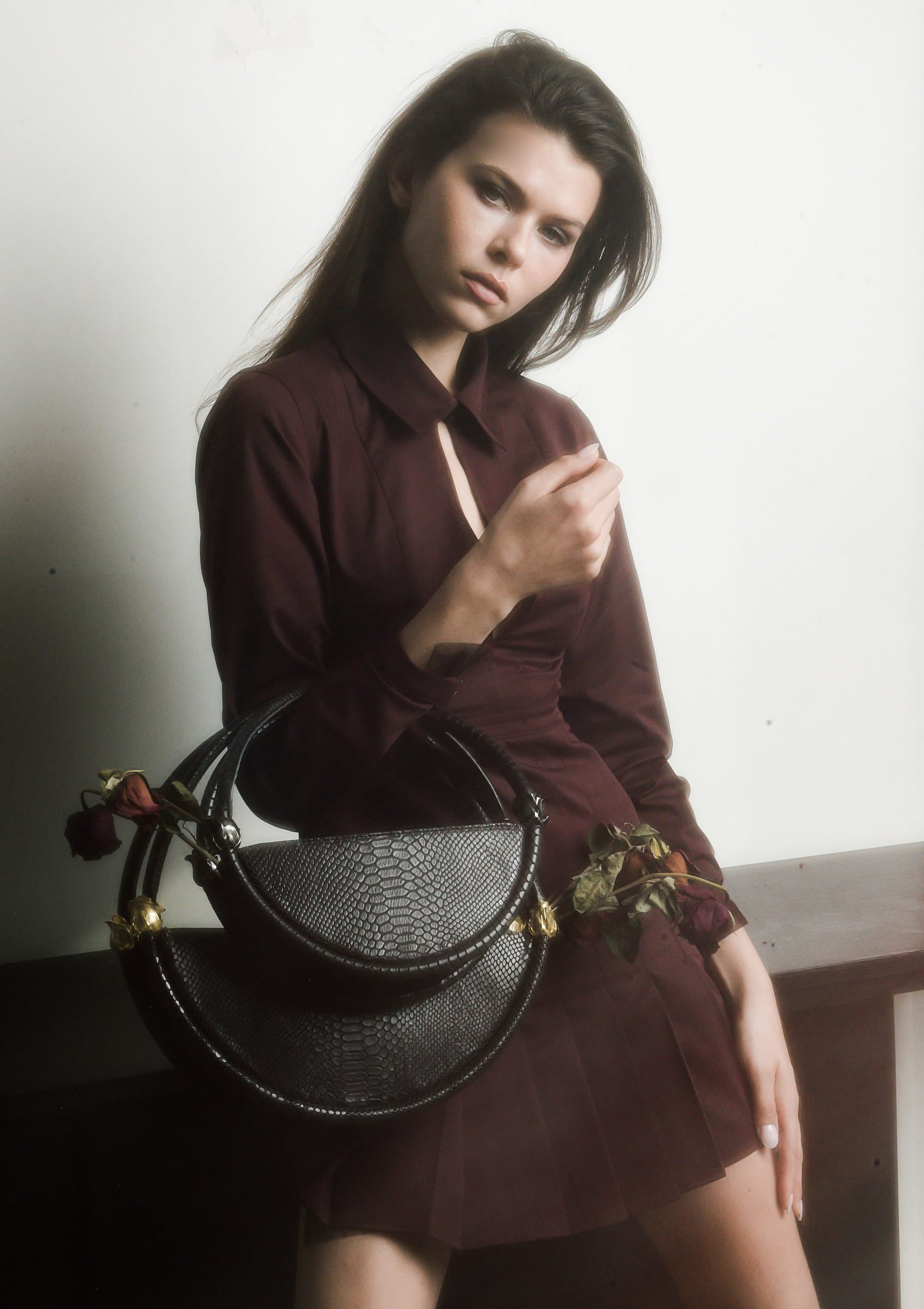 Model wearing a dark burgundy dress holding a textured black handbag against a neutral background.