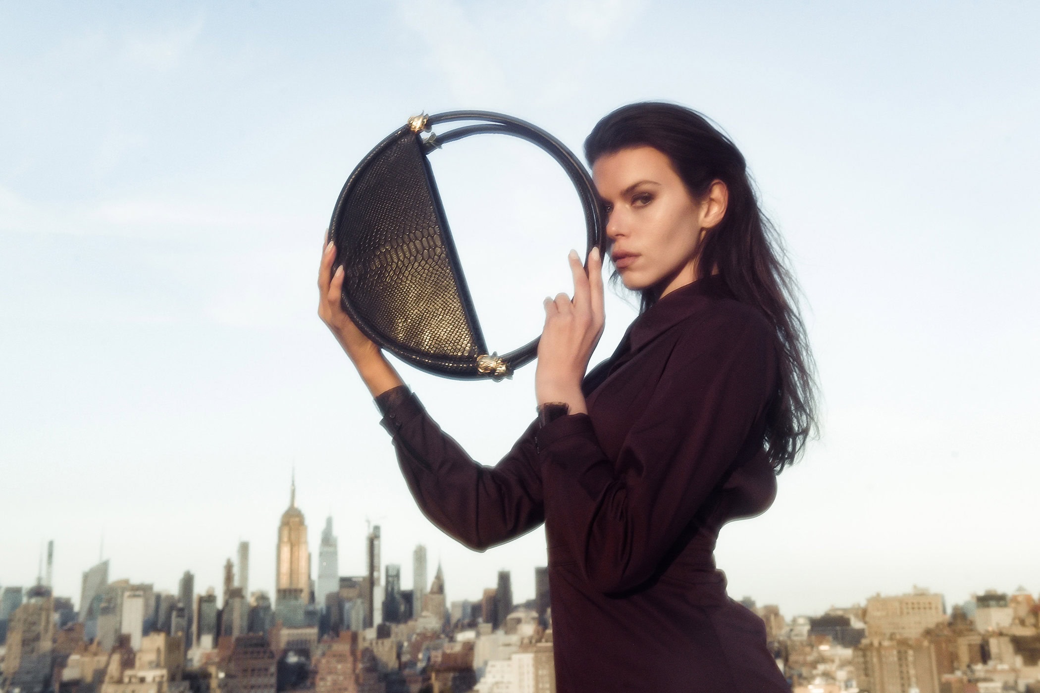 Model holding a round black structured handbag against an urban rooftop skyline backdrop during daylight.