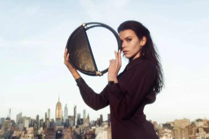 Model holding a round black structured handbag against an urban rooftop skyline backdrop during daylight.