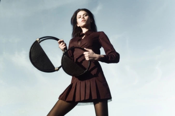 Model Georgia Fowler holding two round black handbags against a clear pale sky for Contessa Mills World Bag 2024 campaign
