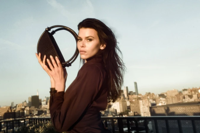 Model holding a dark handbag against a city skyline rooftop backdrop