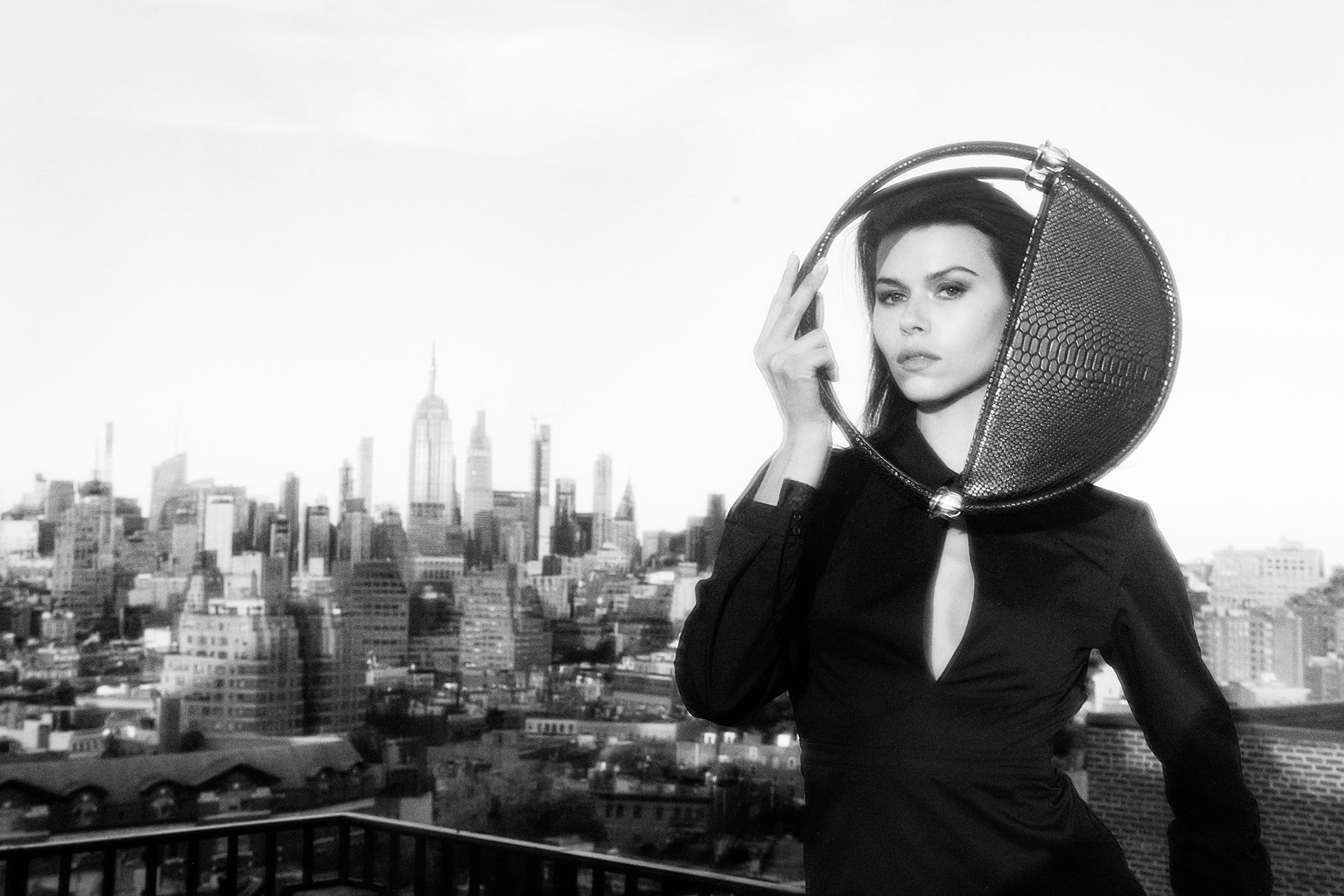 Model holding a round structured handbag on a rooftop overlooking a city skyline in a black and white fashion campaign photograph.
