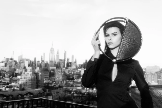 Model holding a round structured handbag on a rooftop overlooking a city skyline in a black and white fashion campaign photograph.