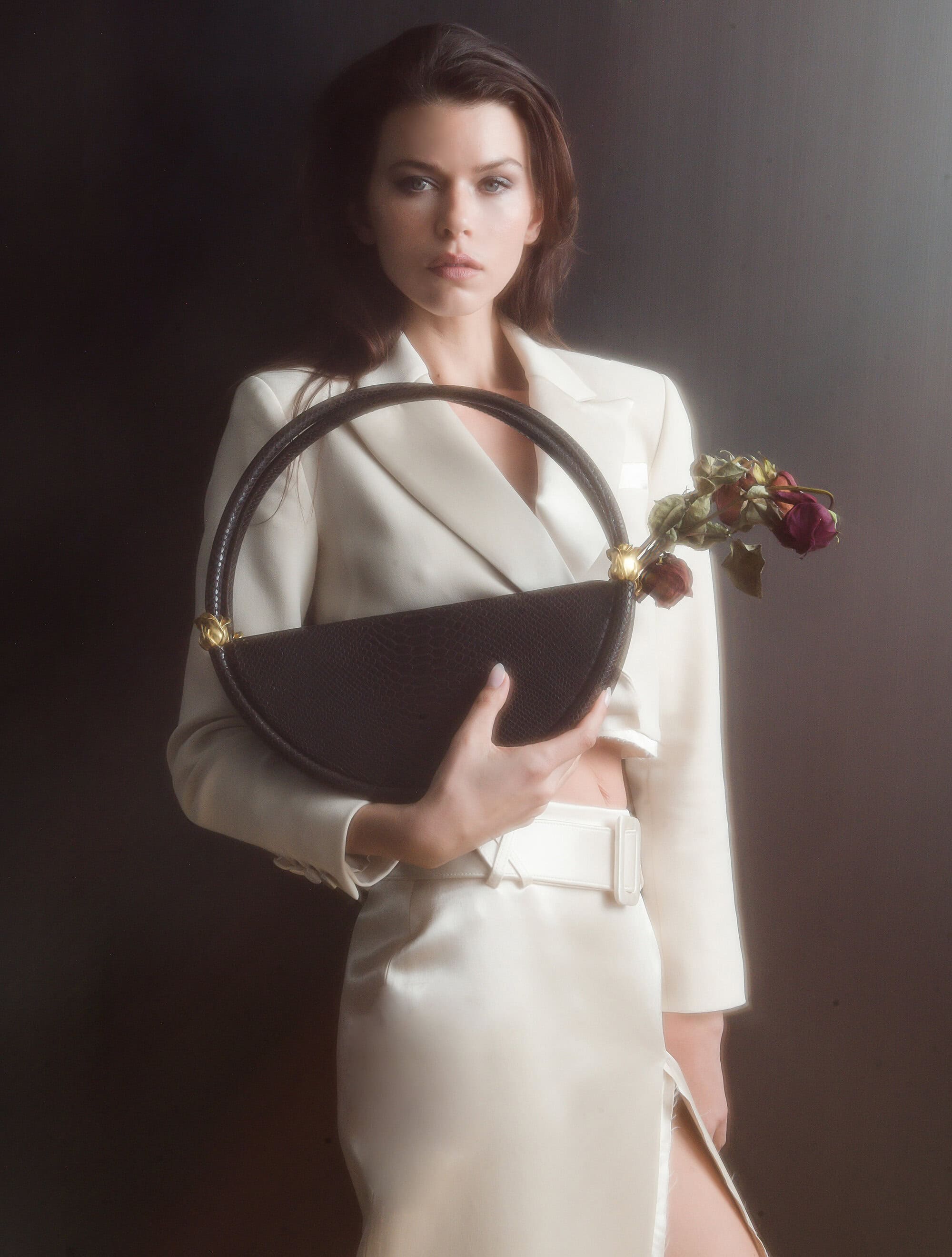 Model wearing a white suit jacket and skirt holding a round black handbag with dried roses for Contessa Mills World Bag campaign