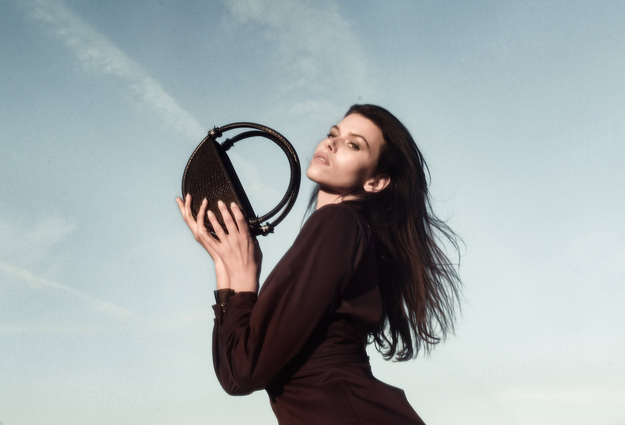 Model Georgia Fowler holding a round black handbag against a pale blue sky in an ad campaign for Contessa Mills by Jacques Burga.
