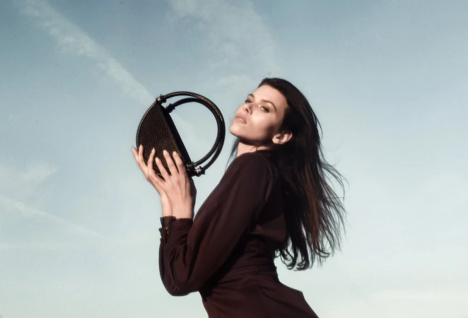 Model Georgia Fowler holding a round black handbag against a pale blue sky in an ad campaign for Contessa Mills by Jacques Burga.