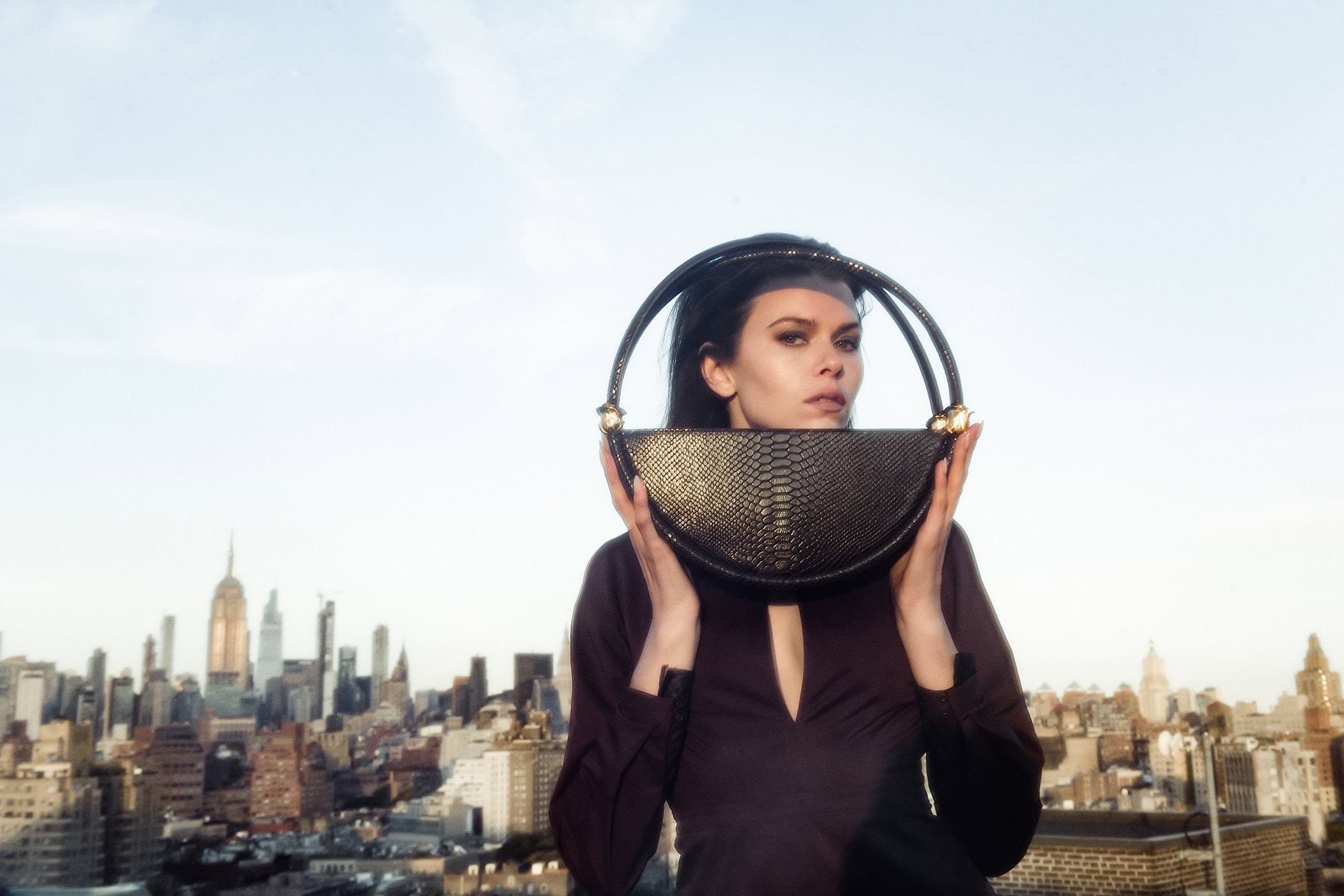 Model holding a round black textured handbag in front of her face against a city skyline backdrop at sunset