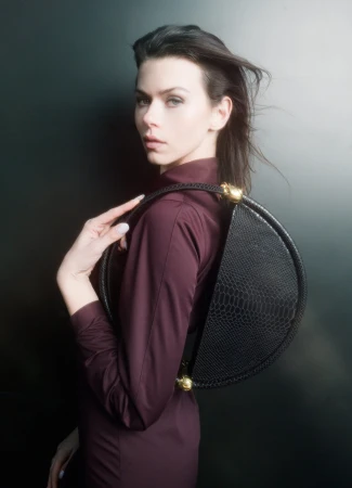 Model wearing a burgundy top holding a circular black handbag against a dark studio background