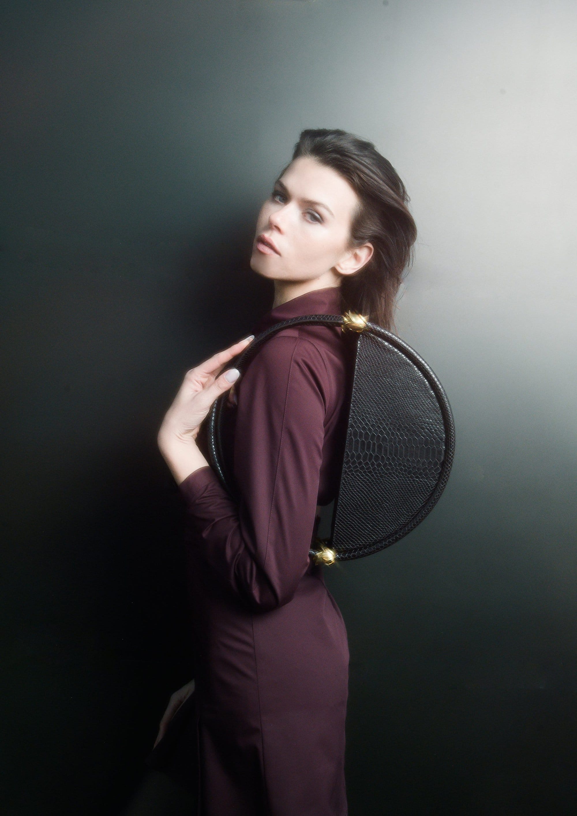 Model Georgia Fowler posing against a dark background holding a round black leather handbag for Contessa Mills World Bag campaign.