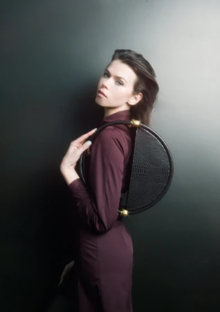 Model Georgia Fowler posing against a dark background holding a round black leather handbag for Contessa Mills World Bag campaign.