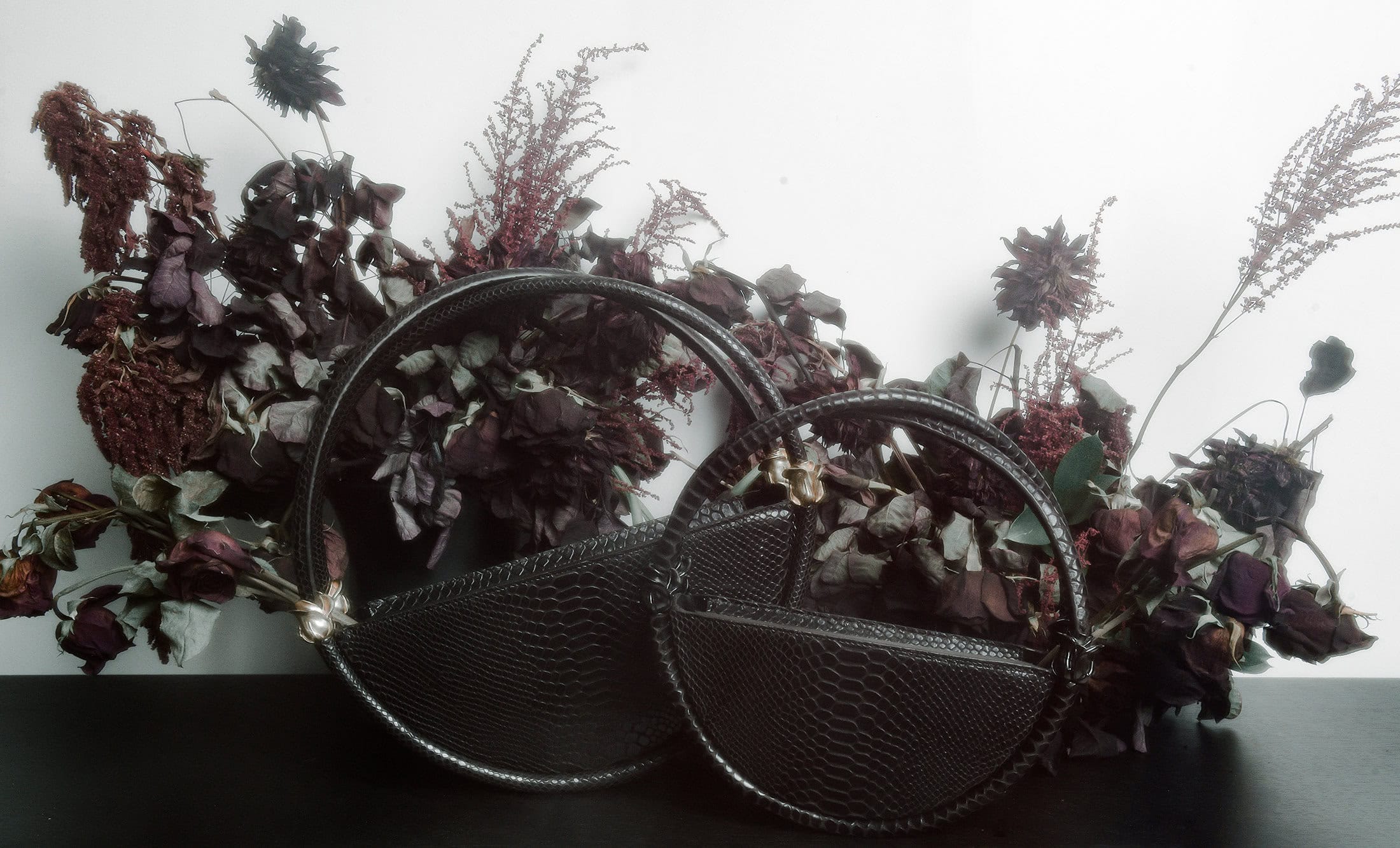 Two black textured round handbags displayed against a dark surface with dark dried flowers and a pale background.
