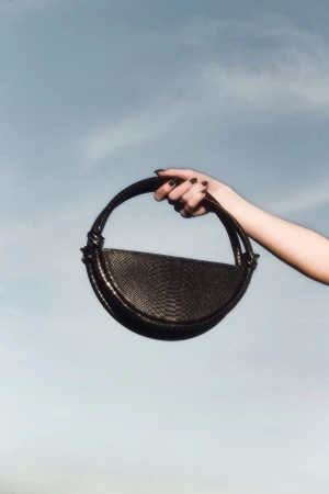 Hand holding a dark crescent-shaped leather handbag against a pale blue sky background