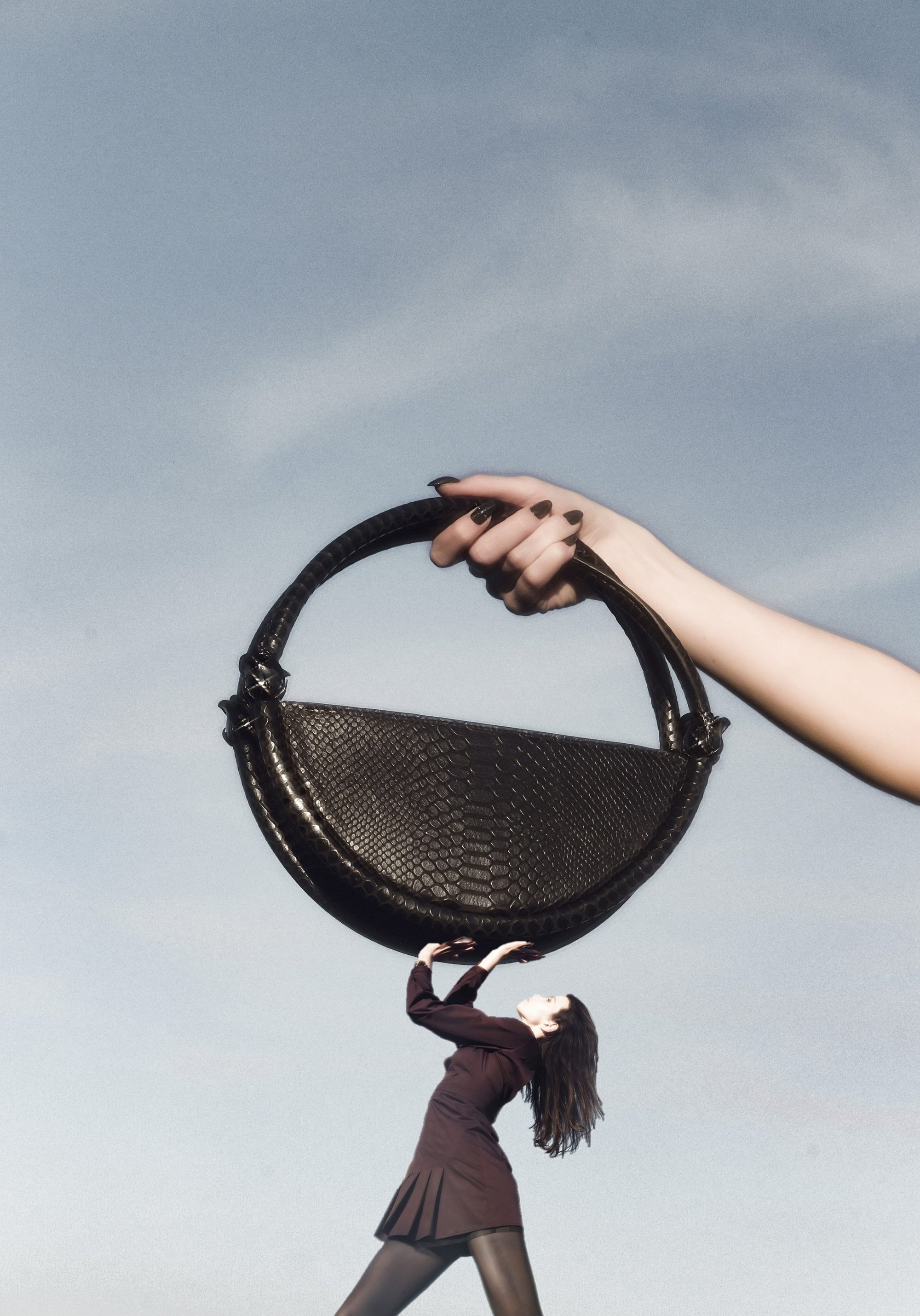 Giant handbag held by a hand from above with a small woman underneath pushing up against a pale sky background