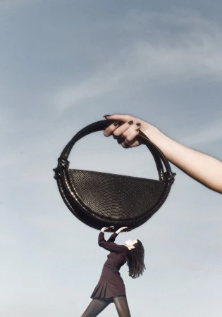 Giant handbag held by a hand from above with a small woman underneath pushing up against a pale sky background