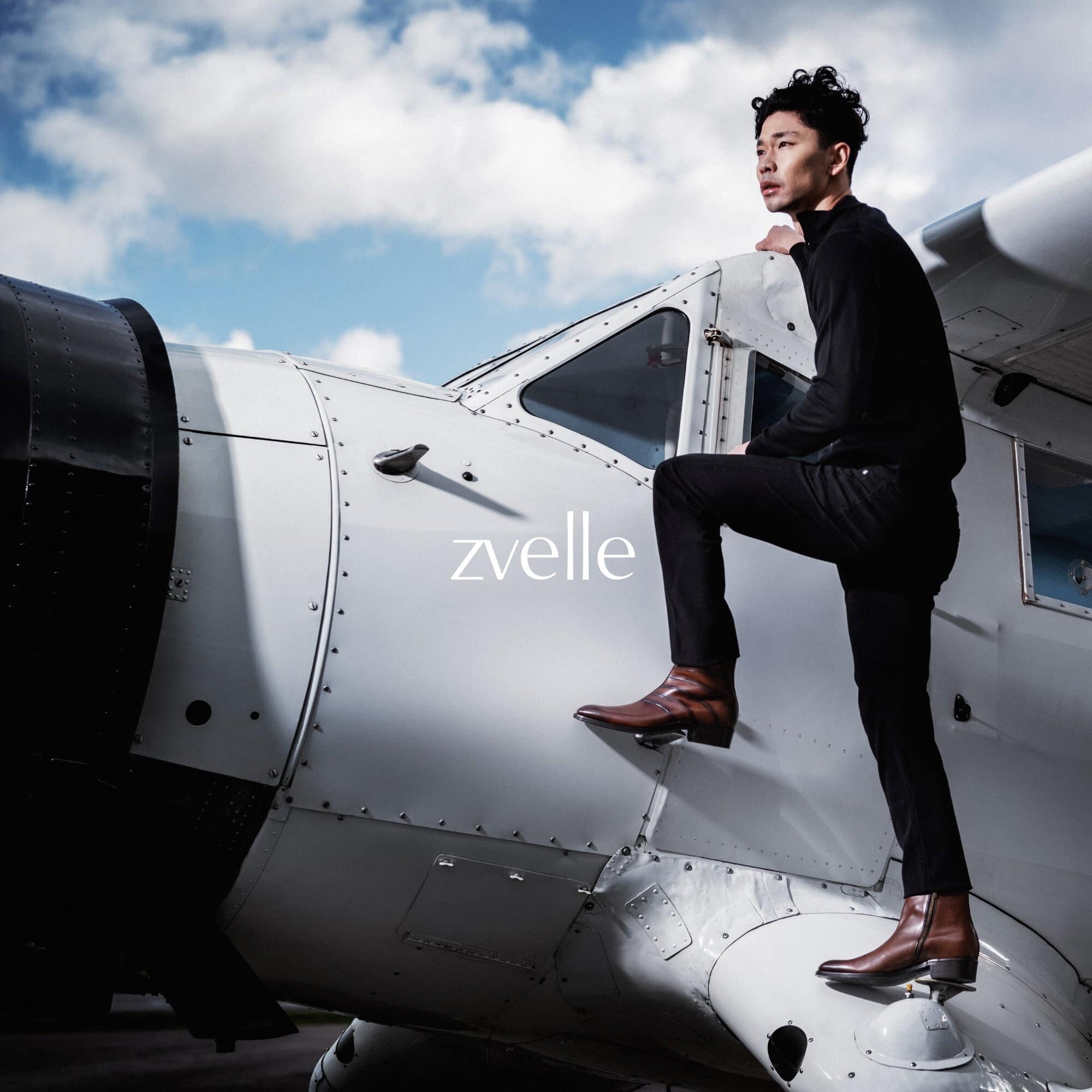 Man wearing black turtleneck, dark pants, and brown leather boots standing on the wing of a vintage airplane
