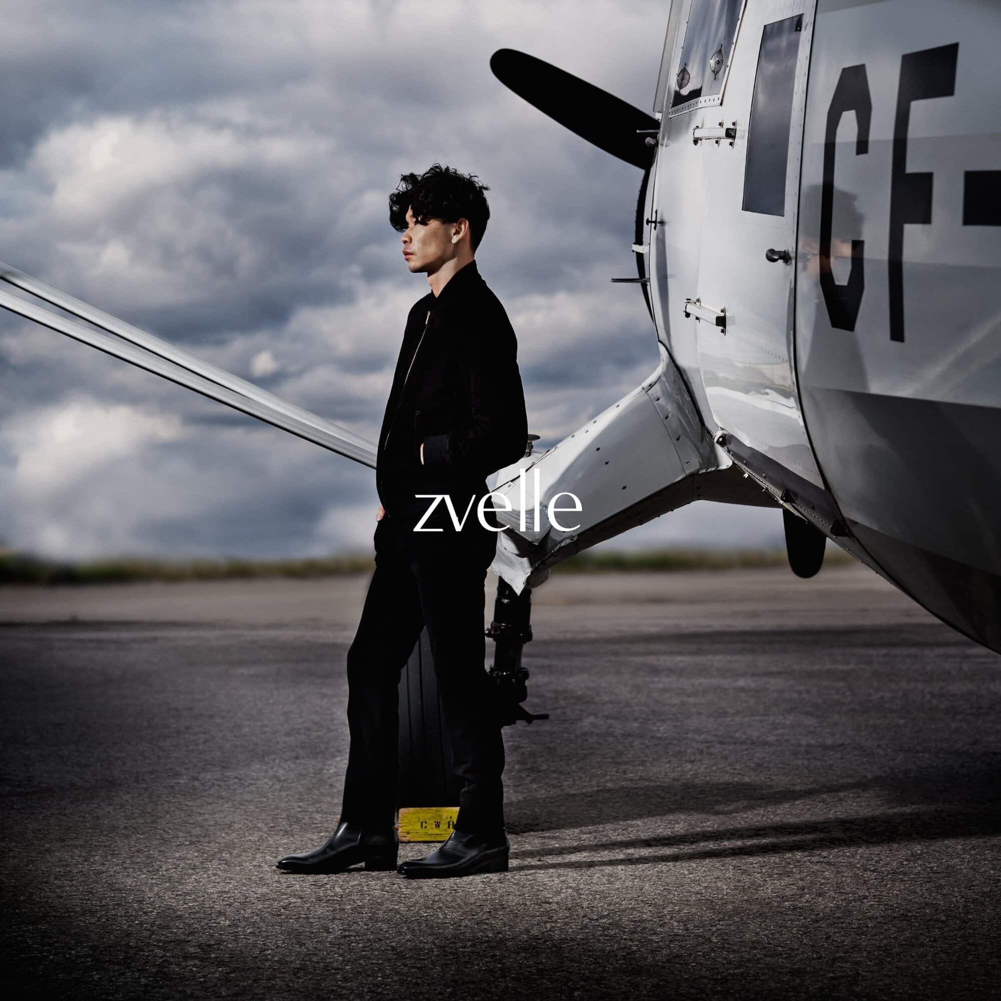 Man standing on an outdoor runway next to a parked vintage airplane, wearing a black jacket and trousers
