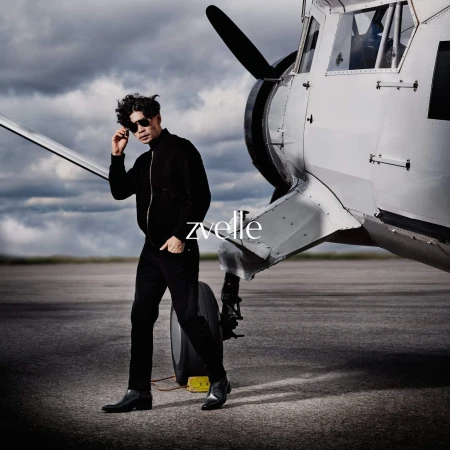 Man wearing sunglasses and black jacket standing in front of a white airplane on an airfield