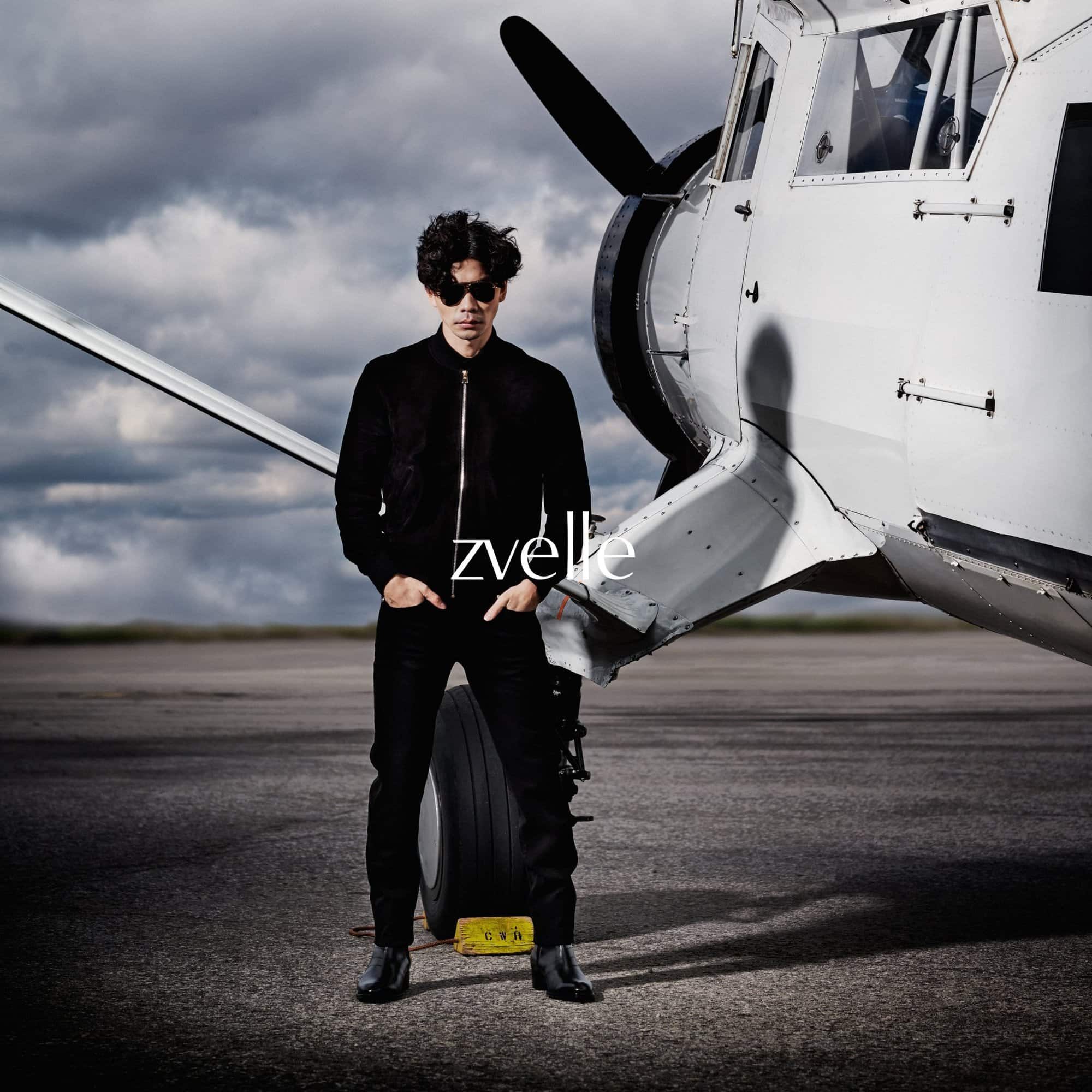 Man wearing dark sunglasses and black jacket standing in front of a white airplane on an airfield