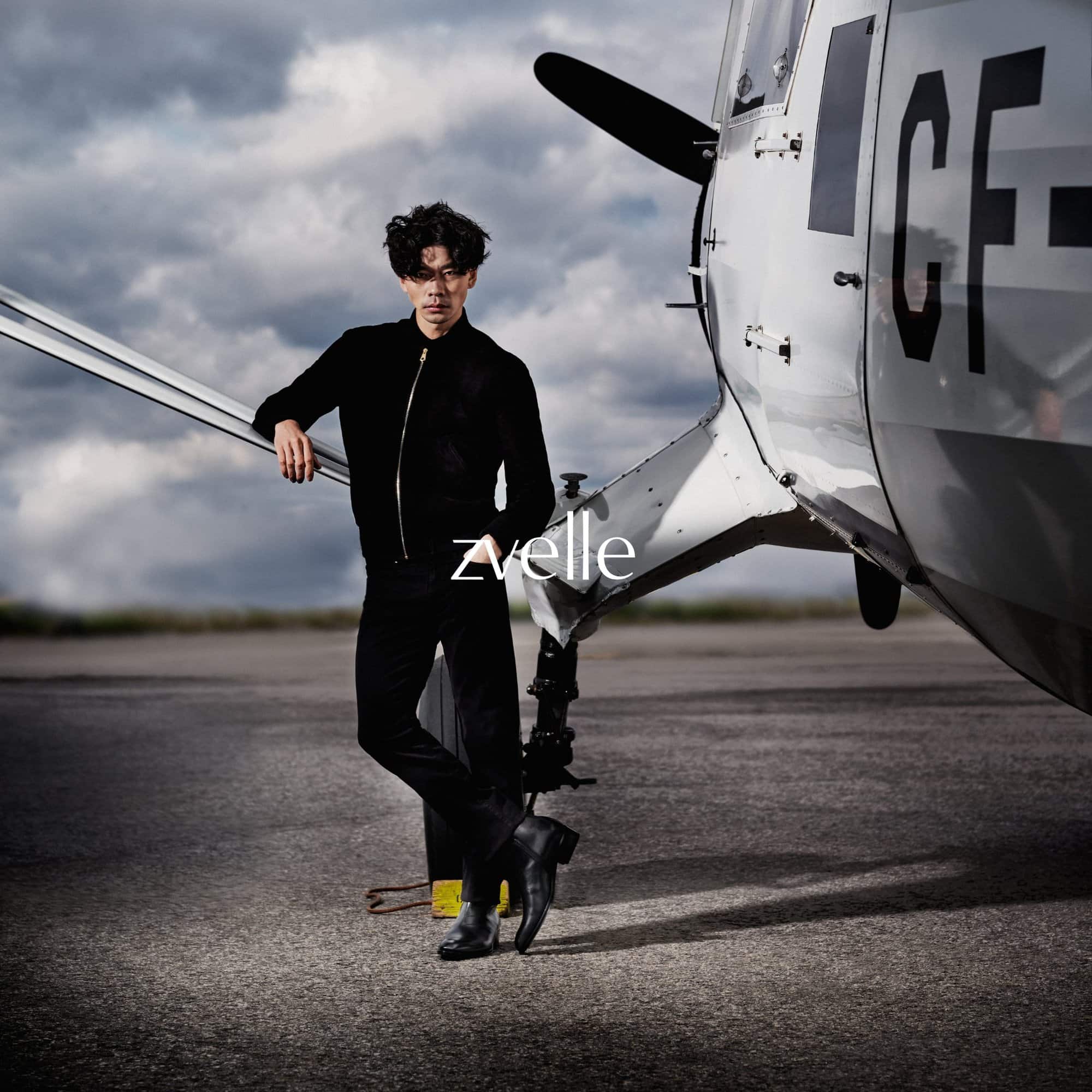 Man in black jacket leaning against a vintage airplane wing on an outdoor airfield under an overcast sky