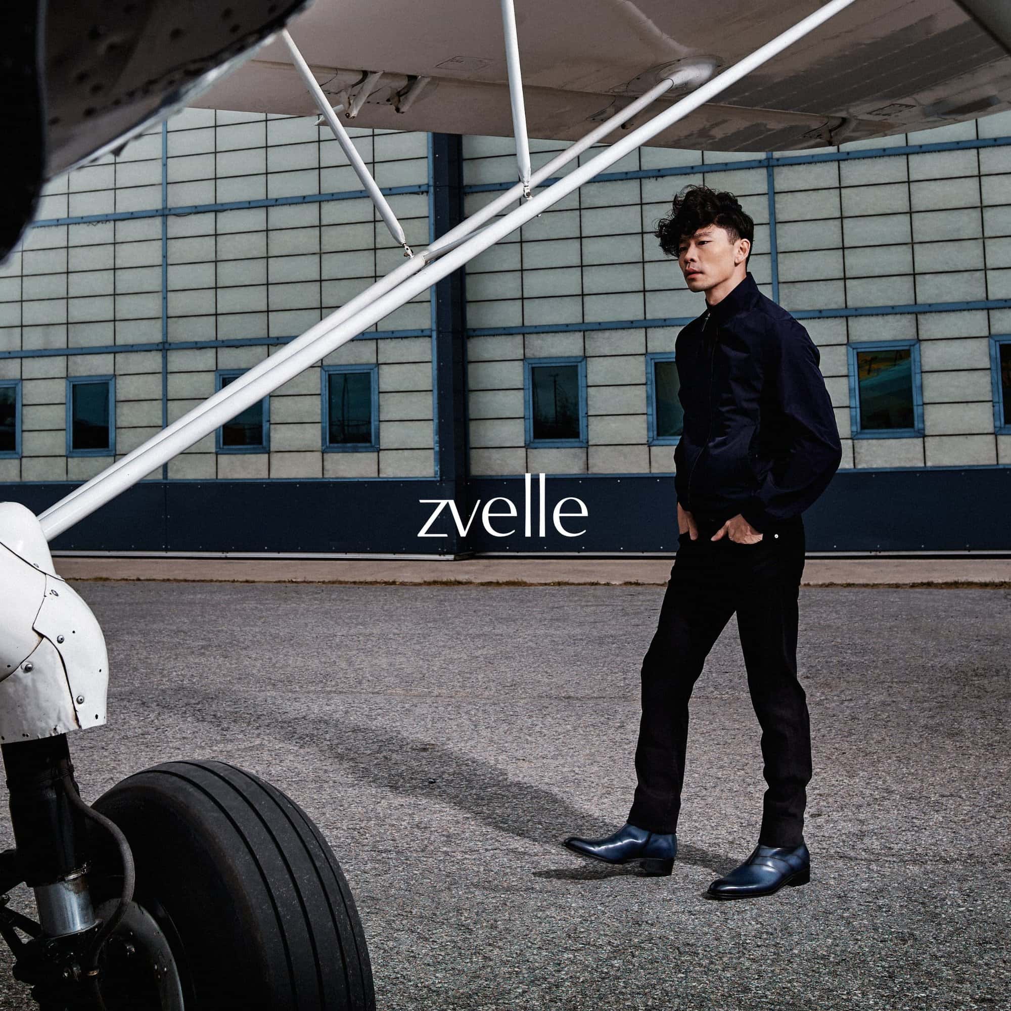 Man wearing dark jacket and trousers standing near an airplane wing and wheel on an airfield