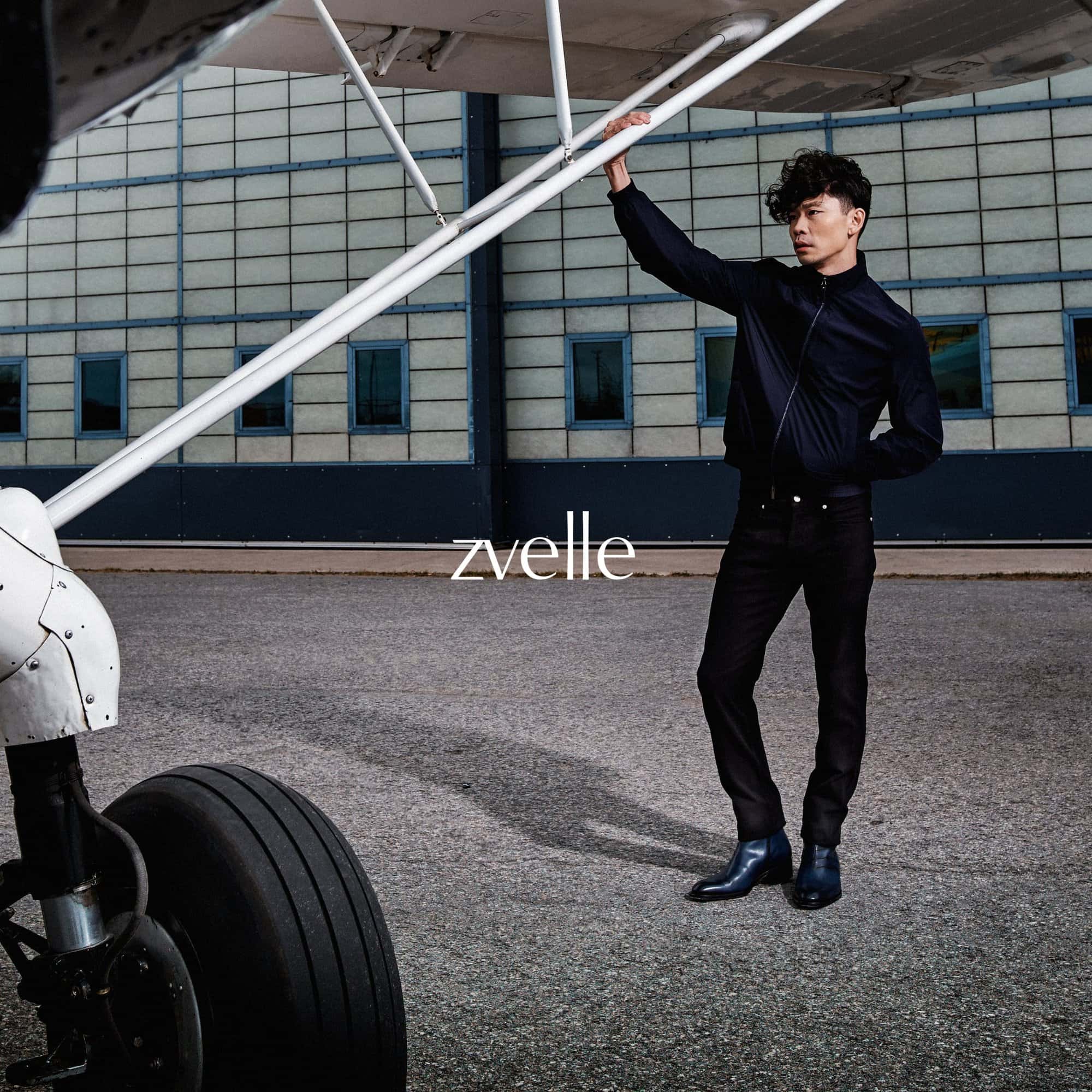 Man wearing a black jacket and trousers standing outdoors next to a small airplane holding a strut