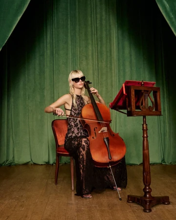 A blond woman wearing sunglasses and a black lace dress seated on a red chair playing a cello on a stage.